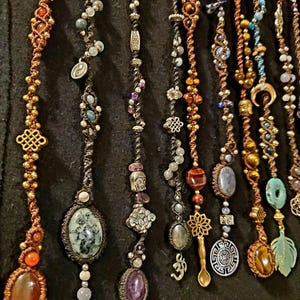 Op de afbeelding: Een verzameling handgemaakte macramé kettingen met diverse edelsteenhangers en decoratieve bedels. De kettingen hebben een verscheidenheid aan kleuren, waaronder bruin, goud, zilver en edelsteentinten. Elke ketting heeft unieke kralen en hangers.