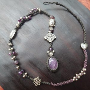 May include: A handcrafted necklace featuring a central oval amethyst stone, accented with silver beads and charms. The necklace includes a Buddha head pendant and a decorative knot design, with a dark cord and purple accents.