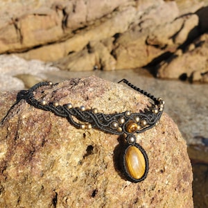 May include: A black macrame choker necklace with gold-colored beads and a large, oval tiger's eye pendant. The necklace is displayed on a textured, brown rock, with a blurred background of rocks and water.