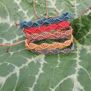 Puede incluir: Una pila de cinco pulseras de macramé con un patrón de nudos trenzados. Las pulseras son de color azul oscuro, rojo, marrón, beige y gris oscuro. Cada pulsera incluye cordones finos ajustables con pequeños nudos en los extremos.