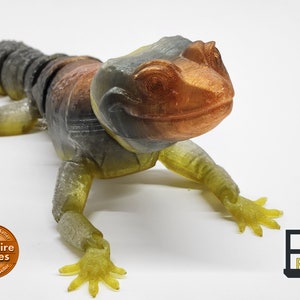May include: A 3D printed lizard figurine with a textured, multi-colored finish. The lizard is in a crawling pose with its head raised and its eyes looking forward. The figurine is printed in a translucent yellow, gray, and brown color scheme. The figurine is printed by MatMire Makes, an authorized seller.