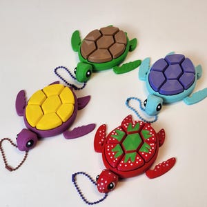 May include: Four colorful turtle keychains with segmented shells. The turtles are purple, green, blue, and red with a green, brown, purple, and green shell respectively. Each turtle has a small chain attached to it.