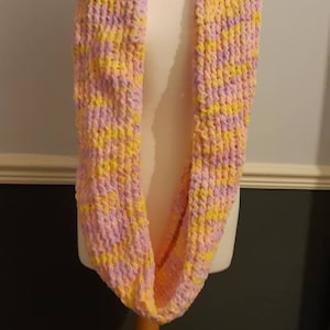 Puede incluir: Bufanda infinita de punto grueso en rosa y amarillo. Se muestra en un maniquí, resaltando su diseño en bucle. El hilo suave y texturizado crea un accesorio acogedor y elegante.
