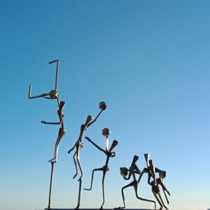 Op de afbeelding: Een metalen sculptuur van zes figuren die basketbal spelen. De figuren zijn gemaakt van spijkers en draad en zijn dynamisch en speels geposeerd. De sculptuur is geplaatst tegen een blauwe lucht achtergrond.