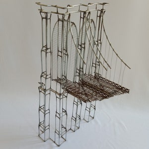 Könnte beinhalten: Eine Draht-Skulptur einer Brücke mit einem Metallgitter-Deck und Drahtseilen. Die Skulptur ist aus silberfarbenem Draht gefertigt und vor einem weißen Hintergrund platziert.
