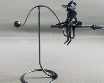 Op de afbeelding: Een metalen sculptuur van een heks die op een bezem rijdt. De heks draagt een puntige hoed en een lange, vloeiende mantel. Het beeld is gemonteerd op een metalen standaard.