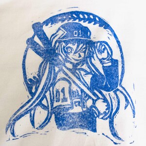 May include: Blue print of an anime-style character with long hair, a hat, and the number "01" on the hat and chest. The character is surrounded by a circular design. The print is on a white background.