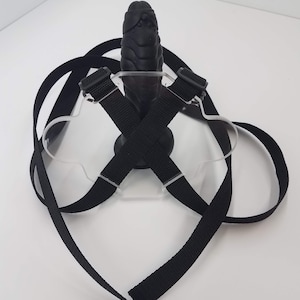 May include: A black harness with straps and buckles, designed to hold a black, textured, rubber object. The harness is made of a clear plastic material.
