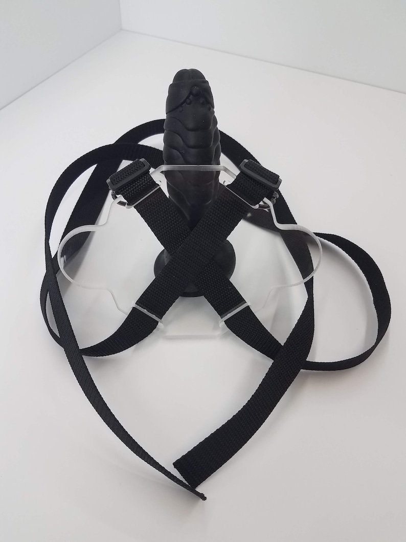 May include: A black harness with straps and buckles, designed to hold a black, textured, rubber object. The harness is made of a clear plastic material.