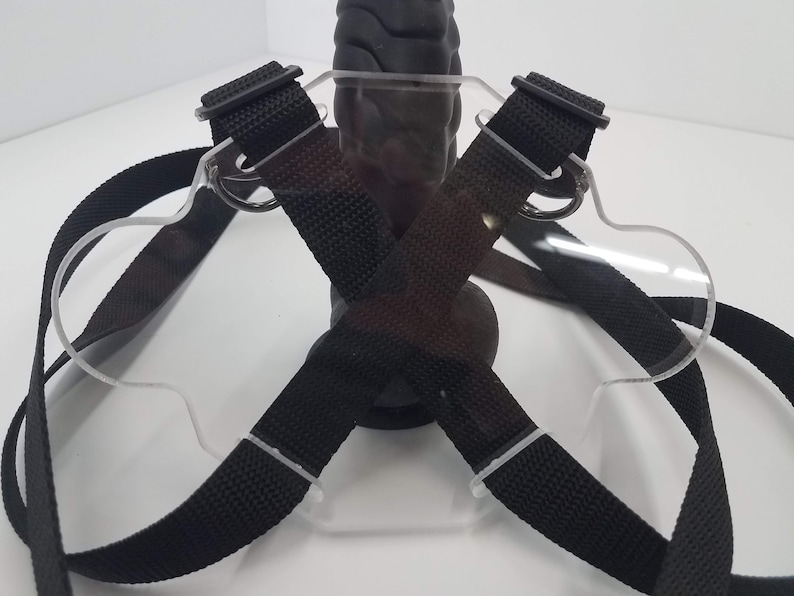 May include: A black harness with clear plastic straps and buckles. The harness is designed to be worn around the waist and chest, and it has a loop in the front for attaching a toy.