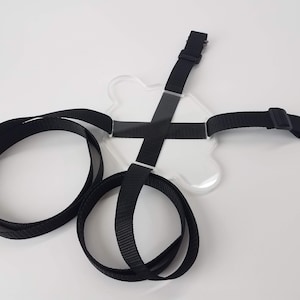 May include: A clear plastic harness with black straps and a metal ring. The harness is designed to hold a phone or other device.
