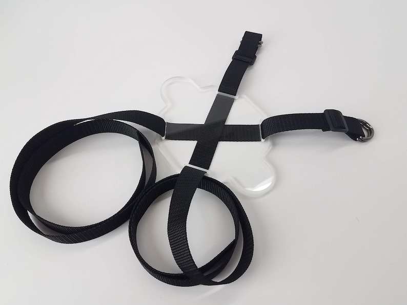 May include: A clear plastic harness with black straps and a metal ring. The harness is designed to hold a phone or other device.
