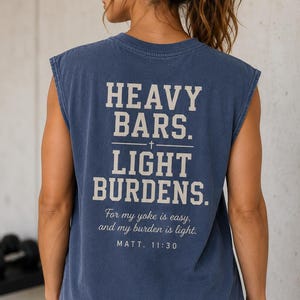 May include: A blue sleeveless t-shirt featuring the text HEAVY BARS. LIGHT BURDENS. with a small cross symbol, followed by the script text For my yoke is easy, and my burden is light. and the citation MATT. 11:30 printed in a light beige color.
