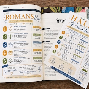 May include: An open Bible featuring colorful tabs and two printed study pages titled The Romans Road and The Hall of Faith. The pages contain numbered lists, religious text, and illustrations in blue, gold, and green, with a Prayer of Surrender.