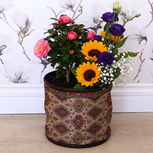 Könnte beinhalten: Ein dekorativer Blumentopf mit einer Vielzahl bunter Blumen. Die Anordnung umfasst rosa Kamelien, gelbe Sonnenblumen, violette Lisianthus und weißen Schleierkraut. Der Topf hat ein gemustertes Stoffdesign.