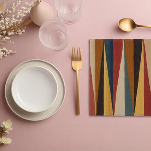 以下が含まれることがあります： 白いボウルとプレート、金色のフォークとスプーン、カラフルな幾何学模様のプレースマットがセットされたテーブルセッティング。透明なグラスと小さなピンクの花瓶も配置され、すべてピンクの表面に配置されています。