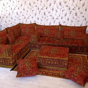 May include: A red and gold patterned sectional sofa set with a matching rug and cushions. The sofa is arranged in an L-shape, with multiple cushions and a low profile. The rug and cushions complement the sofa's design. The set is designed for a living room.