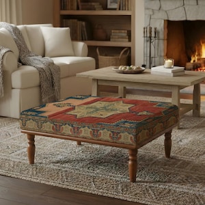 Könnte beinhalten: Eine Wohnzimmerszene mit einem gemusterten Ottoman mit einem rot, blau und goldfarbenen Design. Der Ottoman steht auf einem gemusterten Teppich vor einem weißen Sofa und einem Holztisch. Ein Kamin mit brennendem Feuer befindet sich im Hintergrund.