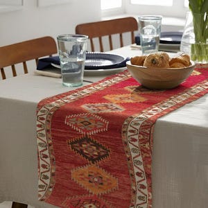 Op de afbeelding: Een rood en oranje tafelloper met een geometrisch patroon, geplaatst op een lichtgekleurd tafelkleed. De loper heeft een gedetailleerde rand en staat op een eettafel met borden, glazen en een houten kom met brood.