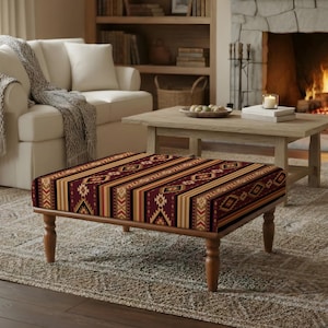 May include: A rectangular ottoman with a patterned top in shades of burgundy, gold, and tan. The design features geometric and striped patterns. The ottoman has a wooden frame and legs, and is placed in a living room setting.