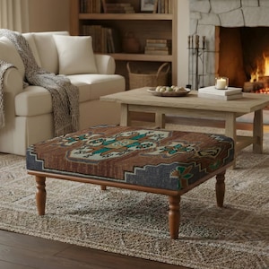 May include: A rectangular ottoman with a patterned top in shades of brown, teal, and blue, set on a wooden frame with four turned legs. The ottoman is placed on a patterned rug in a living room setting, with a white sofa and a fireplace in the background.