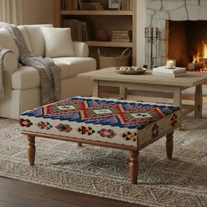 May include: A living room scene featuring a patterned ottoman with wooden legs, a white sofa, and a stone fireplace with a burning fire. A light-colored rug and wooden coffee table complete the cozy setting.