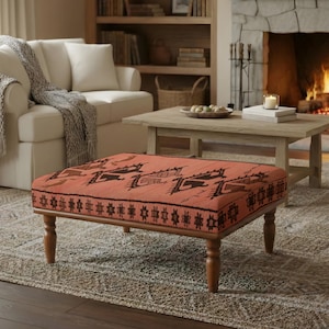 May include: An orange and brown patterned ottoman with wooden legs sits in front of a fireplace. The living room features a white sofa, a wooden coffee table, and a patterned rug, creating a cozy atmosphere.