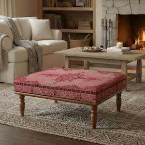 May include: A square ottoman with a pink and red patterned top and wooden legs. The ottoman is in a living room setting with a white sofa, a wooden coffee table, and a fireplace. The rug is beige.