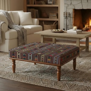 May include: A rectangular ottoman with a patterned top in shades of red, brown, and blue sits on a neutral rug. The ottoman has wooden legs and is in a living room setting with a white sofa, fireplace, and wooden coffee table.
