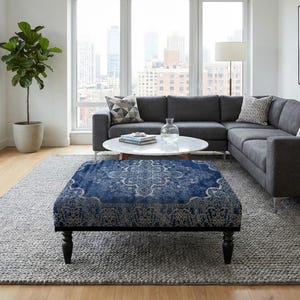 Może przedstawiać: Salon z szarą sofą narożną, okrągłym białym stolikiem kawowym i niebieską pufą we wzory. Duże okno zapewnia naturalne światło. Roślina doniczkowa dodaje zieleni. Pokój jest urządzony w neutralnych tonacjach.