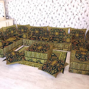 Puede incluir: Un juego de sofás seccionales con una alfombra y cojines a juego. Los muebles presentan un patrón ornamental negro y dorado sobre un fondo verde. El conjunto está dispuesto en un salón con una pared blanca y papel pintado floral.