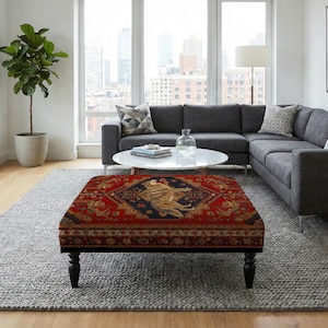 May include: A square ottoman with a detailed, ornate rug top. The rug features a red background with a central design in gold and navy blue. The ottoman has black legs and sits on a gray rug.