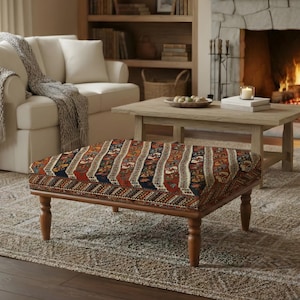 May include: An upholstered ottoman with a patterned top in shades of red, brown, and blue sits on a patterned rug. The ottoman has a wooden frame and legs. A white sofa and a wooden coffee table are in the background.