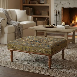 May include: A square ottoman with a patterned fabric top in shades of green, gold, and blue, set on a wooden frame with turned legs. The ottoman is in a living room setting with a sofa, rug, and fireplace.