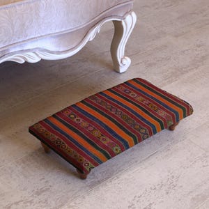 May include: A small, rectangular footstool with a colourful striped pattern in shades of red, orange, and blue. The footstool has a wooden frame and is positioned on a light-coloured floor near a beige upholstered chair.