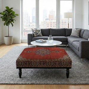 May include: A living room scene featuring a dark gray sectional sofa, a round white coffee table, and a red ottoman with an ornate pattern. A large gray rug and a potted plant complete the decor.