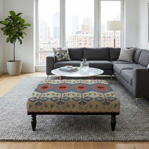 Może przedstawiać: Salon z szarą sofą narożną, wzorzystą pufą i szarym dywanem. Pufa ma beżowe tło z niebieskimi, czerwonymi i zielonymi geometrycznymi wzorami. Obecna jest również duża roślina doniczkowa i okrągły stolik kawowy.