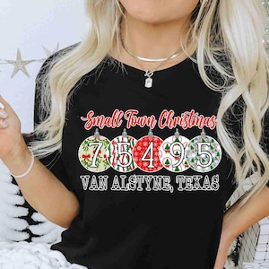 May include: Black t-shirt with the text "Small Town Christmas" above a series of Christmas ornament designs. Each ornament displays a number: 7, 5, 4, 9, and 5. The text "VAN ALSTYNE, TEXAS" is printed below the ornaments.