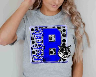 May include: A heather gray t-shirt featuring a large blue letter B with the word Bobcats written vertically in blue script. The design includes a black and white polka dot background pattern and a graphic illustration of a bobcat head.