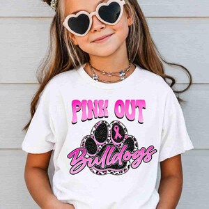 May include: A white short-sleeve t-shirt featuring a graphic print with the text PINK OUT and Bulldogs in pink and black. The design includes a large animal paw print with a pink ribbon symbol and a leopard print pattern in pink and black.