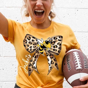 May include: Mustard yellow t-shirt featuring a large leopard print bow with a cartoon bee and the word "Jackets" in script. A brown and white football is held in the right hand.