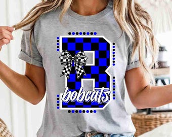 May include: A heather gray short-sleeve t-shirt featuring a large letter B with a blue and black checkered pattern. A black and white checkered bow is attached to the letter, and the word bobcats is written in white script below it.