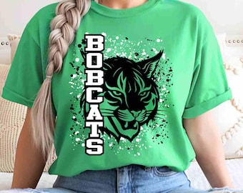 May include: A bright green short-sleeve t-shirt featuring a black graphic of a bobcat head and the vertical text BOBCATS in white block letters with a black outline, set against a background of white and black paint splatter effects.
