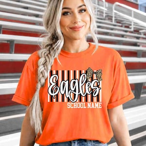May include: An orange short-sleeved t-shirt featuring a graphic print with vertical black and white stripes, the word "Eagles" in white script, a leopard print bow, and the text "SCHOOL NAME" in white block letters centered on the chest.
