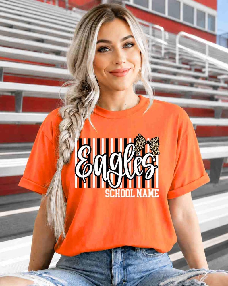 May include: An orange short-sleeved t-shirt featuring a graphic print with vertical black and white stripes, the word "Eagles" in white script, a leopard print bow, and the text "SCHOOL NAME" in white block letters centered on the chest.