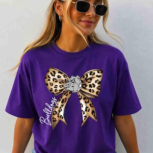 Puede incluir: Una camiseta morada de manga corta con un gr&aacute;fico grande de un lazo con estampado de leopardo centrado en el pecho. Una peque&ntilde;a ilustraci&oacute;n de un bulldog gris est&aacute; colocada en el centro del lazo y la palabra Bulldogs est&aacute; escrita en letra cursiva blanca.