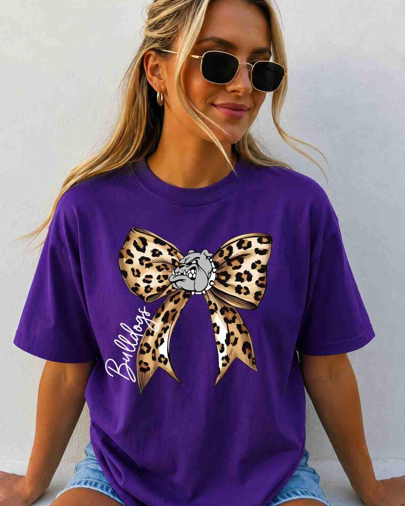 Puede incluir: Una camiseta morada de manga corta con un gr&aacute;fico grande de un lazo con estampado de leopardo centrado en el pecho. Una peque&ntilde;a ilustraci&oacute;n de un bulldog gris est&aacute; colocada en el centro del lazo y la palabra Bulldogs est&aacute; escrita en letra cursiva blanca.