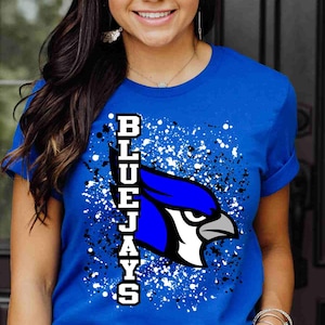 May include: A royal blue short-sleeved t-shirt featuring a graphic of a blue jay bird head in profile. The design includes the vertical text "BLUEJAYS" in white block letters with a black outline, set against a background of black and white paint splatters.