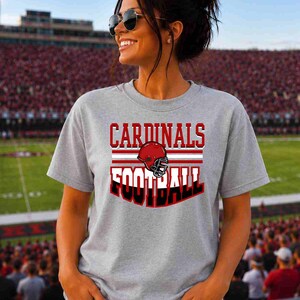 May include: A heather gray short-sleeve t-shirt featuring the text CARDINALS FOOTBALL in red and black lettering, centered above and below a graphic of a red football helmet with a black face mask and white horizontal stripes.
