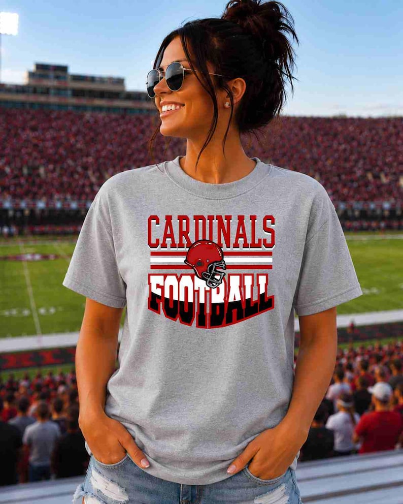 May include: A heather gray short-sleeve t-shirt featuring the text CARDINALS FOOTBALL in red and black lettering, centered above and below a graphic of a red football helmet with a black face mask and white horizontal stripes.